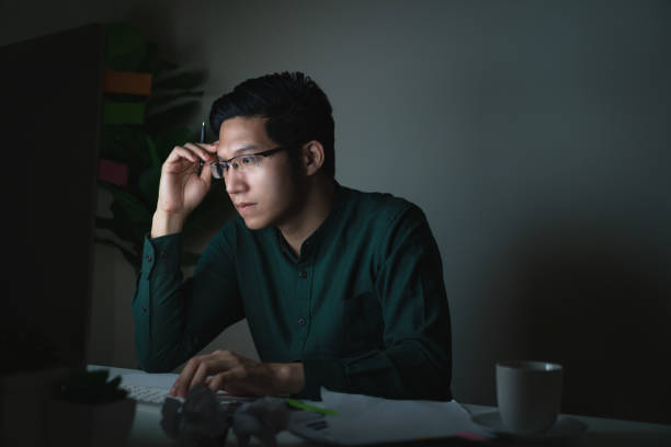 attraente giovane asiatico seduto sul tavolo della scrivania a guardare il computer portatile al buio a tarda notte lavorando sentendosi serio pensando e determinato a casa in ufficio nel lavoro duro o nel concetto di lavoro carico di lavoro. - working late working computer busy foto e immagini stock