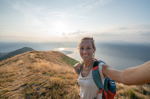 Cheerful young woman taking a selfie portrait on mountain top after hiking all the way up reaching summit