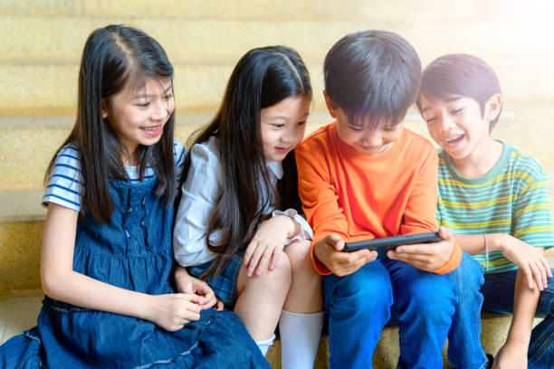 cute little asian children using mobile phone, playing games, watching video movie together while sitting on steps outdoors - homework teenager mobile phone school imagens e fotografias de stock