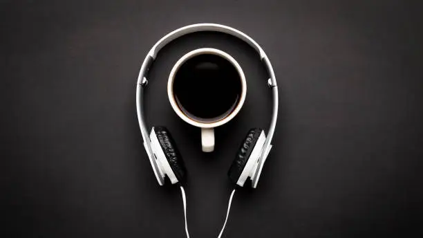 Coffee in white mug and white headphones on a black background. Top view, copy space