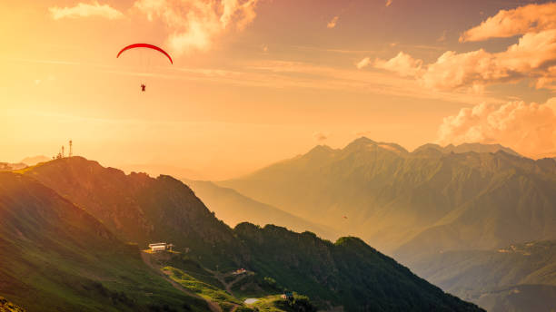 roten gleitschirm in den orangefarbenen sonnenuntergang bewölkten himmel über die grünen berge. grünen tal mit seilbahn nach unten unten. krasnaja poljana, sotschi, kaukasus, russland. - paragliding sport austria parachuting stock-fotos und bilder