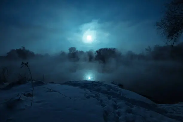 Photo of Night mystical scenery. Full moon over foggy river. Snow covered riverbank.