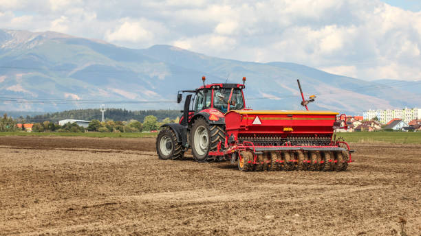 istock Red tractor pulling sowing trailer behind over empty field. Some village and mountains in background. 1090056048