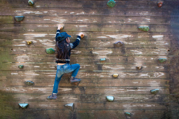 Children play outdoors on a wooden playground in a park. Happy girl playing on wooden climbing wall in playground during summer.