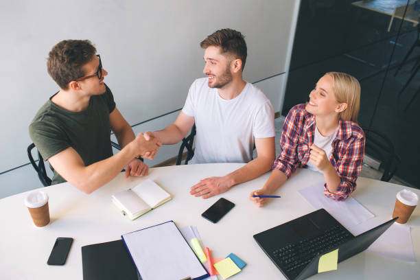 Two nice and cheerful guys sit in front one another and shake hands. They smile. Young blonde woman look at them and laugh. They are together in one room. Two nice and cheerful guys sit in front one another and shake hands. They smile. Young blonde woman look at them and laugh. They are together in one room incubator stock pictures, royalty-free photos & images