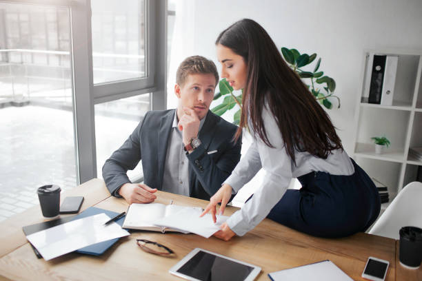 istock Young beautiful woman sit on table and point on document. She looks down. Guy hold hand on mouth. 1090086920