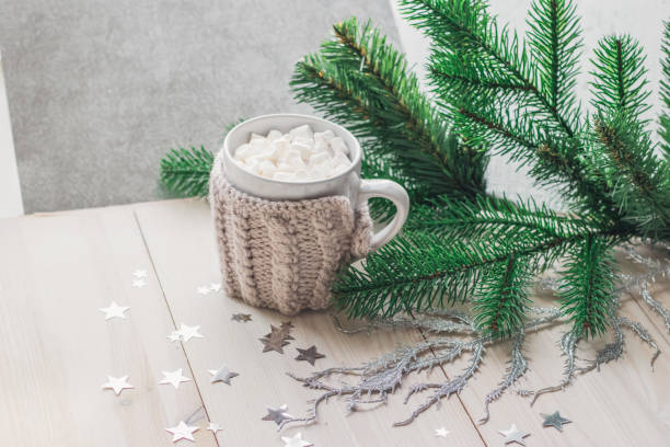 istock White cup with marshmallows in a warm knitted jacket on a light wooden background with a Christmas tree and silvery stars. 1090106700