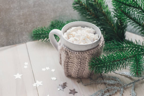 istock White cup with marshmallows in a warm knitted jacket on a light wooden background with a Christmas tree and silvery stars. 1090106924
