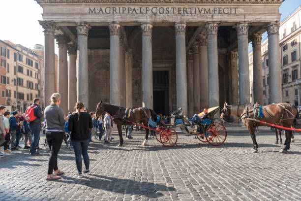 Rome, Italy - April 8, 2017: A wide angle image of the Piazza della Rotunda with a horse-drawn carriage (parked in the street) and tourists (and possibly some local Romans) on a morning in spring. A ride with a horse drawn carriage is a popular way to explore central Rome. The image was captured just in front of the Pantheon. The Pantheon ('to every god') in Rome, Italy was commissioned by Marcus Agrippa as a temple to all the gods of Ancient Rome, and built by Emperor Hadrian in about 126 AD. The inscription on the wall translates to: “Marcus Agrippa, son of Lucius, made this building during his third consulship”. The piazza (city square) was made on the order of Pope Eugenius IV (1431–1439). It took its name from the Pantheon, which had been converted into the Christian church (informally) known as Santa Maria Rotonda (in the 7th century AD). In the center of the piazza is a fountain, the Fontana del Pantheon (constructed by Giacomo Della Porta under Pope Gregory XIII in 1575), surmounted by an Egyptian obelisk (added to the fountain in 1711 under Pope Clement XI).