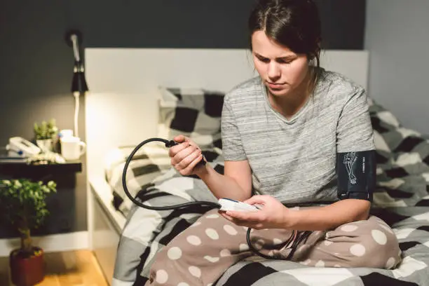 Photo of The topic of high blood pressure is hypertension disease. 22 year old young Caucasian woman uses an automatic tonometer to check pressure at home in the bedroom on the bed in home clothes in evening