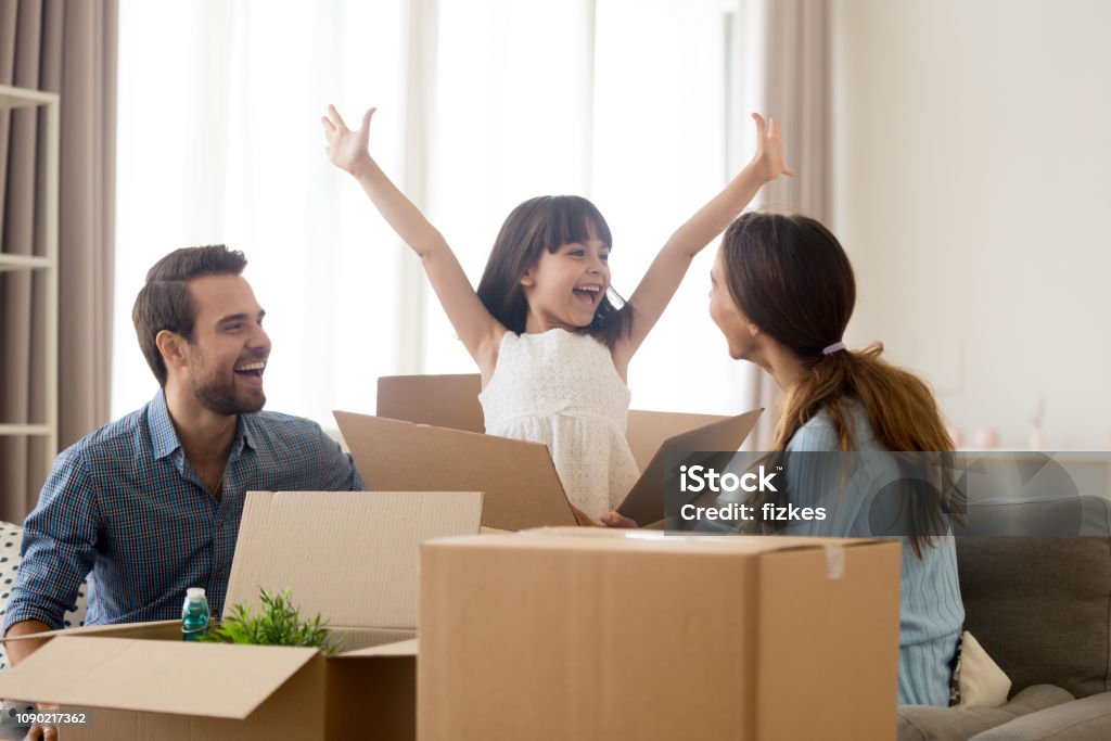 Happy kid jumping out of box laughing packing with parents Happy kid daughter jumping out of box excited about moving day or relocation, cheerful child girl playing unpacking in new home with mom and dad, smiling family laughing having fun packing together Family Stock Photo