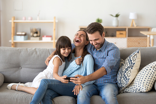 Cheerful family vloggers with child daughter taking selfie, recording vlog, making video call on phone having fun together in living room, parents and kid girl laugh using cellphone funny online app