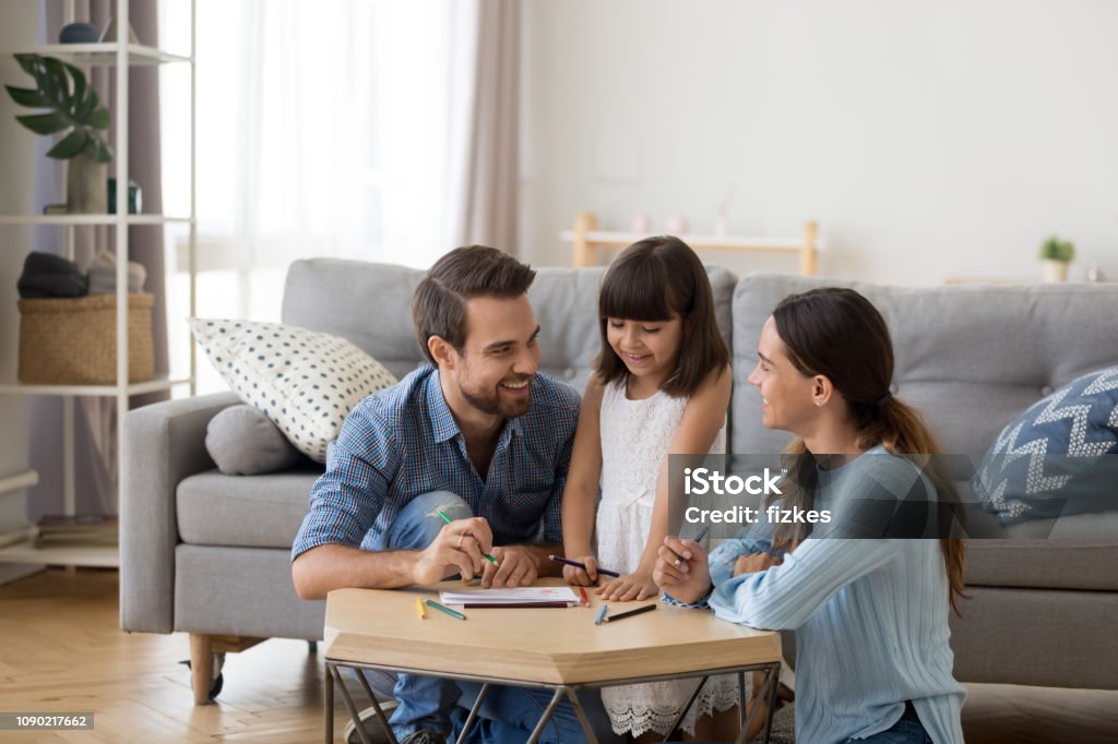 Caring mom and dad teaching little daughter to draw Happy family with kid playing together, caring mom and dad smiling teaching little daughter to draw with color pencils, mother and father having fun with cute child help in creative weekend activity Family Stock Photo