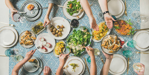 Flat-lay of human hands holding mediterranean starters and drinks Mediterranean style family or friends dinner. Flat-lay of table with various salads, starters and pastries over blue table cloth with human hands holding glasses with drinks and sharing food, top view. Holiday gathering, celebration, vegetarian party concept restaurant table layout stock pictures, royalty-free photos & images