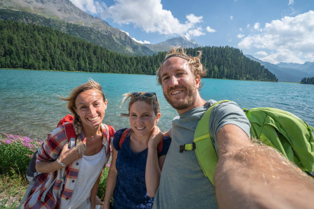 tres jóvenes de senderismo en verano y tomando un retrato selfie por un bellísimo lago cordillerano - silvaplanersee fotografías e imágenes de stock