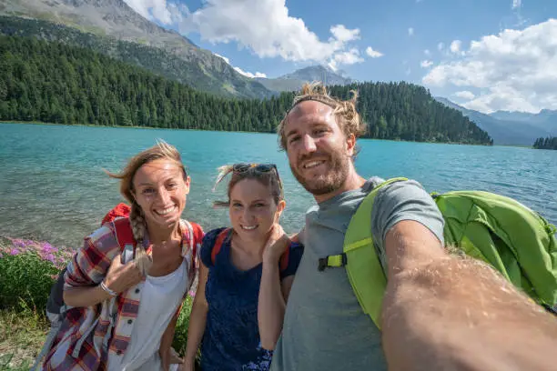Three young people hiking in Summer and taking a selfie portrait by a beautiful mountain lake in Switzerland, Graubunden Canton