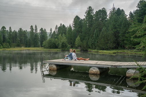 Cheerful young couple sitting on lake pier and sharing some relaxing moments.\nPeople travel nature concept