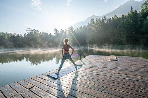 One young woman exercising yoga on a lake pier in the morning, fog on water surface. People wellbeing relaxation healthy lifestyle concept.\nShot in Graubunden Canton, Switzerland.