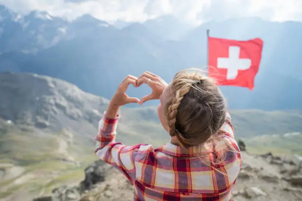 Young woman making heart shape finger frame on mountain top overlooking the Swiss Alps; Swiss flag