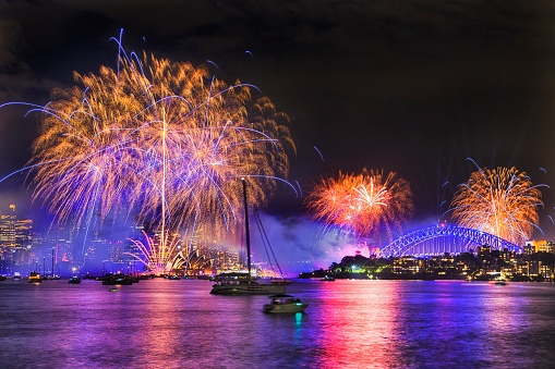 Calligraphic drawing in dark midnight sky of Sydney city during New year fireworks with splashes of colourful light balls over harbour water, CBD towers and Harbour bridge arch.