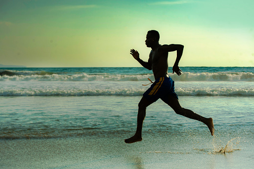 silhouette of young attractive fit athletic and strong black African American man running at sunset beach training hard and sprinting on sea water in professional athlete and runner workout