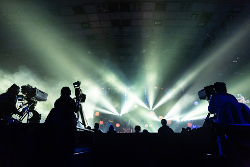 Silhouette of a group of cameramen broadcasting an event. Workers are on a high platform on the background of bright beams.
