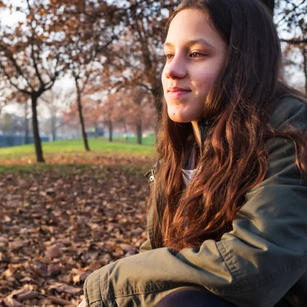 Portrait of pretty teenage girl with brown long hair and green jacket, autumn background with trees and foliage, copy space for text