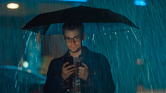 Young Caucasian Man in Glasses, Wearing a Jeans Coat and Square Shirt is Using a Smartphone Under an Umbrella. It's Dark Outside and it is Raining.