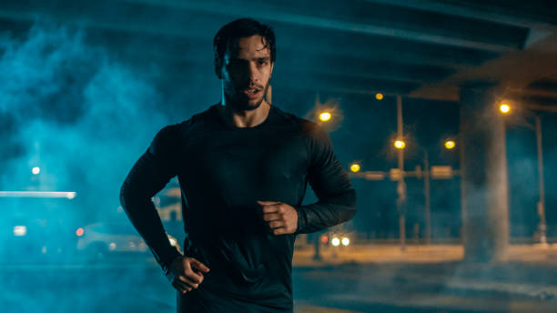 sweating tired athletic muscular young man in sports outfit jogging in a street filled with smoke. he is running in an evening urban environment under a bridge. - night running imagens e fotografias de stock