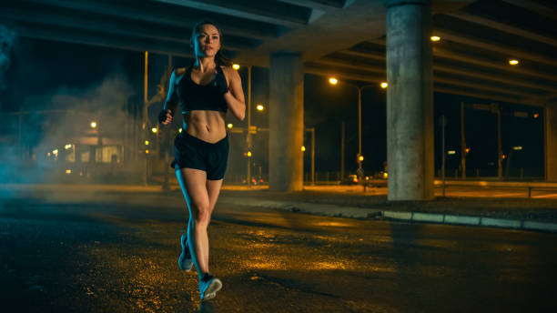 beautiful fitness girl in black athletic top and shorts is jogging on the street. she is doing a workout in an evening wet urban environment under a bridge. - night running imagens e fotografias de stock