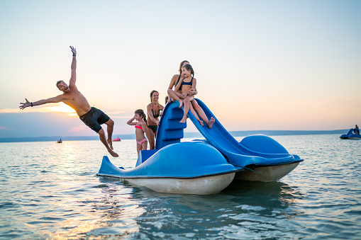 family sister children girls boy having fun with paddleboat padel boat with slide on lake in summer sunset series funny father jumping in water