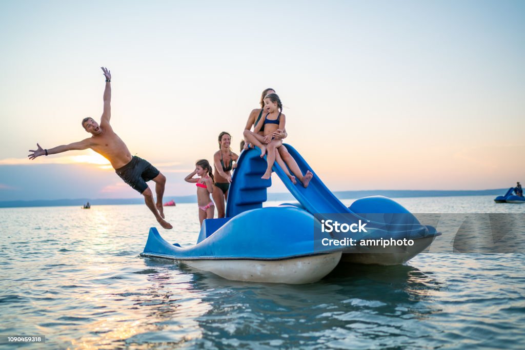 happy family enjoying summer sunset on lake with pedal boat family sister children girls boy having fun with paddleboat padel boat with slide on lake in summer sunset series funny father jumping in water Pedal Boat Stock Photo