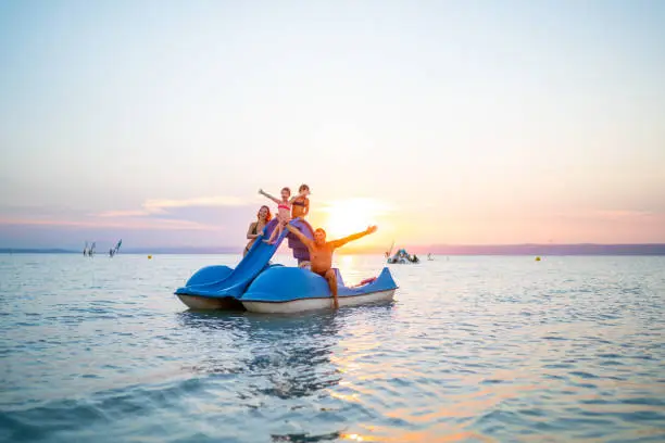happy family with two children girls having fun with paddleboat pedal boat with slide on lake in summer sunset series