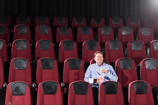 A mature man in his 50s, sitting in a movie theater alone, surrounded by empty seats. He is watching the movie, eating popcorn.