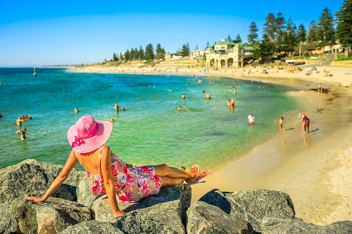 Woman in hat looking at the turquoise waters of Cottesloe Beach, Perth's most famous town beach, Western Australia. Girl sunbathing on the rocks of the Indian Ocean. Summer season with blue sky.