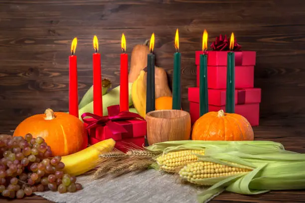 Photo of Kwanzaa holiday concept with candles red, black and green, gift box, pumpkins, ears of wheat, grapes, corns, banana, bowl and fruits on wooden background, close up