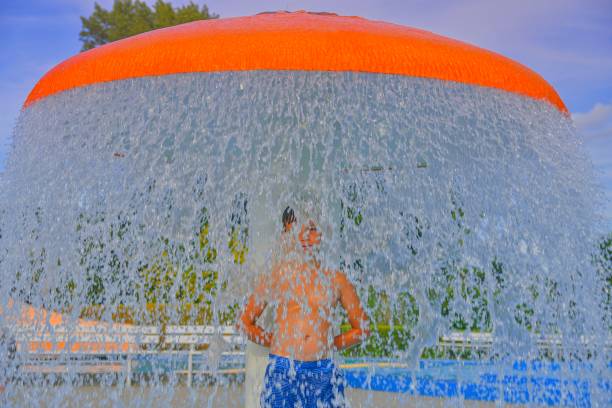 Happy little boy enjoying summer day in the swimming pool. Boy standing under sprinkler in spray pool. Summer and happy chilhood concept stock photo