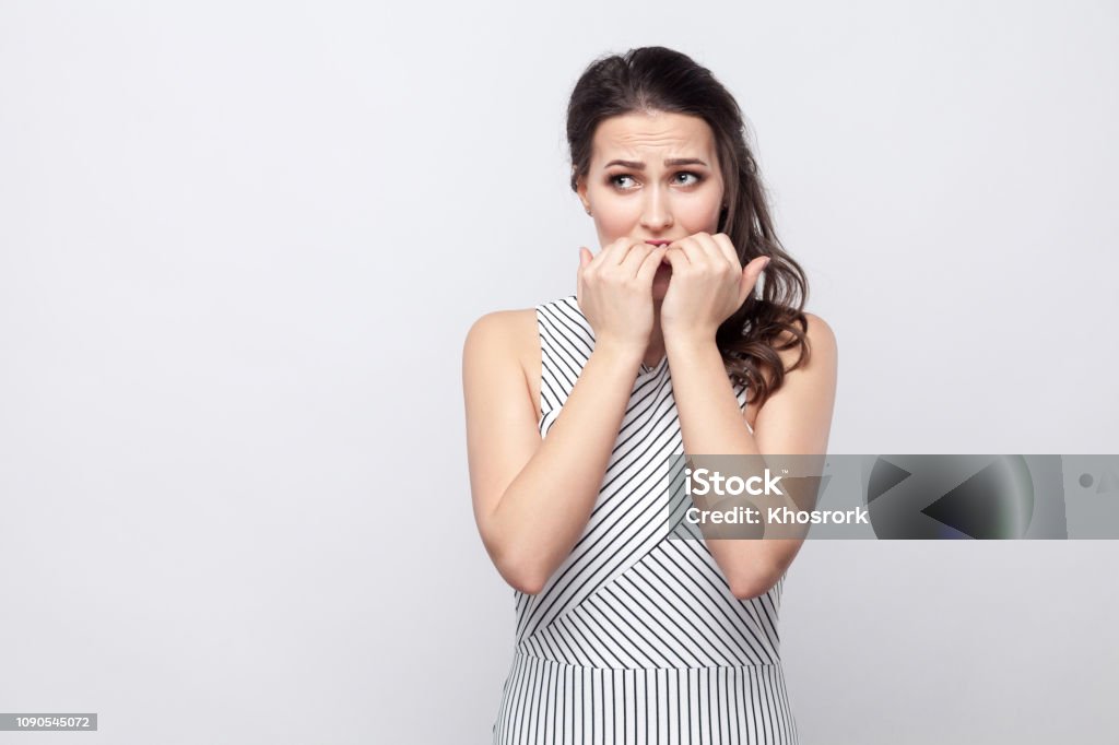 Portrait of worry beautiful young brunette woman with striped dress standing, looking away and bitting her nails with stressed face Portrait of worry beautiful young brunette woman with striped dress standing, looking away and bitting her nails with stressed face . indoor studio shot, isolated on grey background. Nail Biting Stock Photo