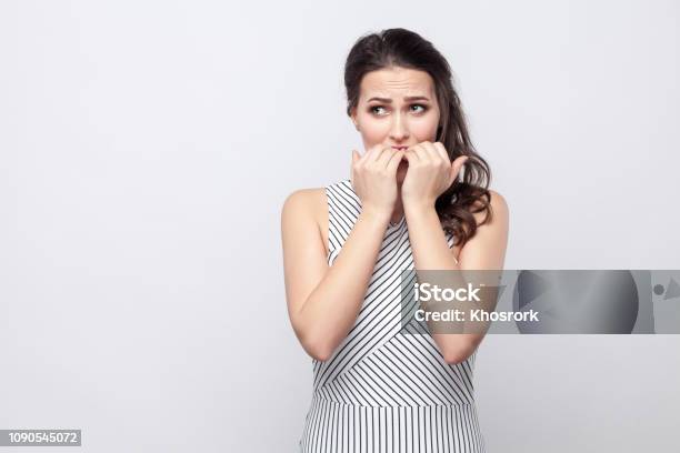 Portrait Of Worry Beautiful Young Brunette Woman With Striped Dress Standing Looking Away And Bitting Her Nails With Stressed Face Stock Photo - Download Image Now