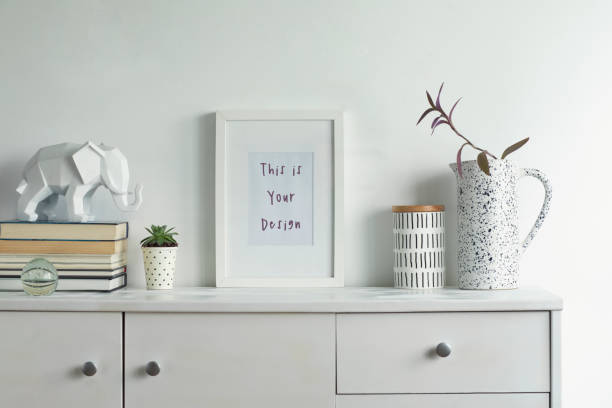 Scandinavian stylish cupboard with mock up poster frame, elephant figure, design vase, accessories, books, cacti. White background wall.