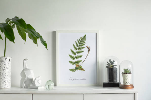 Scandinavian modern cupboard with mock up poster frame, design vase, tropical leaf, succulents in glass pot and cat figure. White background wall in minimalistic interior.