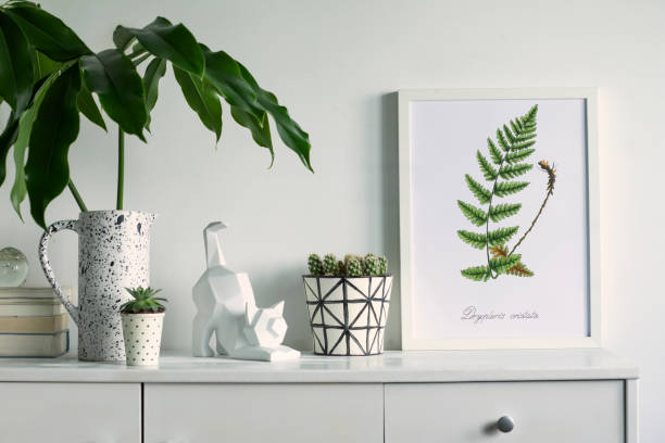 istock Scandinavian stylish cupboard with mock up poster frame, cat figure, tropical leaf, accessories, books, cacti. White background wall. 1090546788