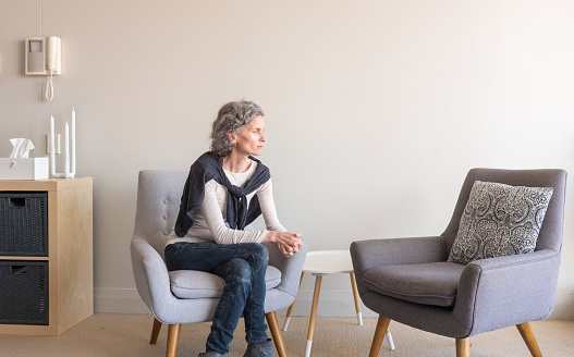 Middle aged woman with grey hair seated alone in living room with empty armchair (selective focus)