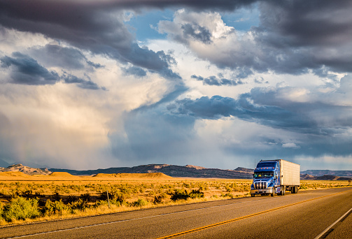 Beautiful panorama view of classic semi trailer truck on empty highway with dramatic sky in golden evening light at sunset