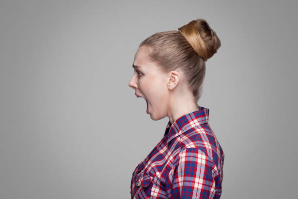 profile side view of scared blonde girl in red, pink checkered shirt, collected bun hairstyle standing and looking aside and screaming profile side view of scared blonde girl in red, pink checkered shirt, collected bun hairstyle standing and looking aside and screaming. indoor studio shot. isolated on gray background women screaming surprise fear stock pictures, royalty-free photos & images