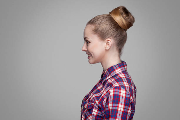 profile side view of beautiful blonde girl in red, pink checkered shirt, collected bun hairstyle standing and looking aside with toothy smile. profile side view of beautiful blonde girl in red, pink checkered shirt, collected bun hairstyle standing and looking aside with toothy smile. indoor studio shot. isolated on gray background braided buns stock pictures, royalty-free photos & images
