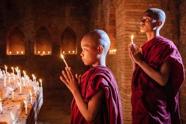 Myanmar Novice Monks Praying at Buddha Statue Two young Burmese buddhist monks in their typical clothing inside temple praying together to worship buddha. Natural Light inside dark temple with giant Buddha Statue. Bagan, Mandalay Region, Myanmar. mandalay photos stock pictures, royalty-free photos & images