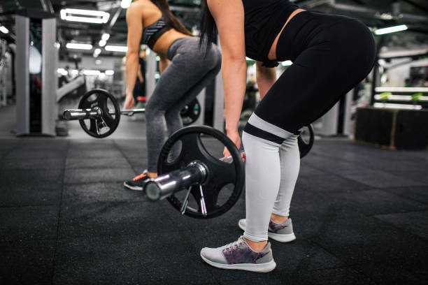 istock Cut view og two young women working with barbell and weight. They squad and lean forward. Models have sexy bodies. 1090772130