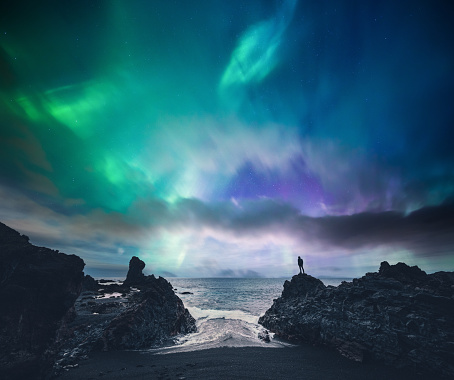 Man standing on the rock at the beach watching northern lights (Iceland).