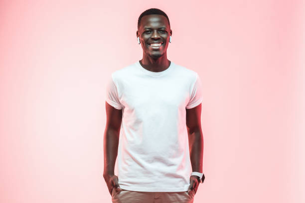 young african american man standing with hands in pockets, wearing blank white t-shirt with copy space for your logo or text, isolated on pink background - artificial model imagens e fotografias de stock
